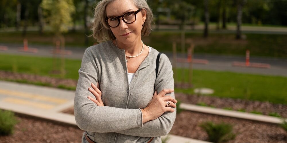 Mature woman with glasses and crossed arms stands confidently outdoors in a park setting during daylight