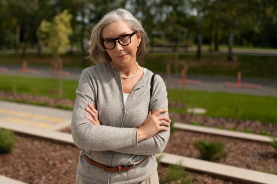 Mature woman with glasses and crossed arms stands confidently outdoors in a park setting during daylight