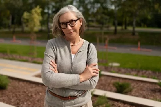 Mature woman with glasses and crossed arms stands confidently outdoors in a park setting during daylight